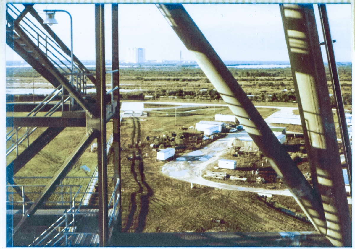 Viewed between the emergency egress stair tower and main-framing structural members on Column Line 7 of the Rotating Service Structure at Space Shuttle Launch Complex 39-B, Kennedy Space Center, Florida, the Apollo-era Vertical Assembly Building and last intact Launch Umbilical Tower can be seen in the far distance standing above the horizon.